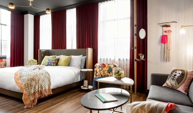 Ένα μεγάλο δωμάτιο με ένα κρεβάτι μπροστά από παράθυρα από το δάπεδο μέχρι την οροφή επενδεδυμένα με πλούσια κόκκινα κουρτίνες και έναν καναπέ και τραπέζι στο πλάι στο The Broadview Hotel στο Τορόντο του Καναδά