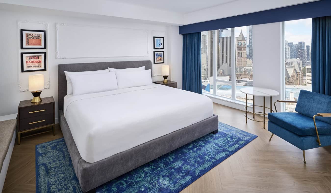 Ένα king-size κρεβάτι με κορνίζες που κρέμονται από τους τοίχους και στις δύο πλευρές και μεγάλα παράθυρα από το δάπεδο μέχρι την οροφή με θέα στο αστικό τοπίο του Τορόντο, Καναδάς, στο Pantages Hotel Downtown